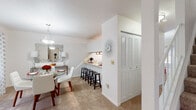 This two-floor residence is designed with a functional and well-connected layout. The living room features plush carpeting and a sliding glass door that opens to an outdoor space, allowing natural light to filter in and providing views of surrounding greenery. The room offers a comfortable, open feel suited for everyday living.

Adjacent to the living area, the dining space is defined by a pendant light fixture overhead and a window dressed with patterned curtains. A pass-through counter connects the dining area directly to the kitchen, where a row of barstools creates an additional casual seating option. The kitchen itself is equipped with standard appliances, warm wood cabinetry, light countertops, and tile flooring.

A set of louvered closet doors near the dining area provides practical storage. The primary bedroom is generously sized with carpet flooring, a large window with natural light, and direct access to a bathroom. An additional room visible in the layout functions as a secondary bedroom or office space. The bathroom features a wide vanity countertop, a large mirror, and clean, neutral finishes throughout. The overall aesthetic is bright, neutral, and cohesive across both floors.