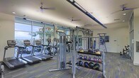 This fitness center offers a comprehensive workout environment designed to accommodate a wide range of exercise routines. The space is anchored by a wall of floor-to-ceiling windows that flood the interior with natural light while providing views of palm trees and the surrounding outdoor environment, creating an energizing atmosphere for exercise.

The cardio zone features multiple treadmills and elliptical machines arranged along the windowed wall, allowing users to enjoy the outdoor scenery during their workouts. Overhead, ceiling fans are evenly distributed throughout the space, ensuring proper air circulation and comfort during intense training sessions.

The functional training area is well-stocked with a multi-station cable and pull-up rig, along with a tiered rack holding an extensive collection of kettlebells and medicine balls in various weights. A large wall mirror enhances the sense of space and allows users to monitor their form. Recessed lighting supplements the natural light, ensuring the space remains well-lit throughout the day. The durable, patterned flooring ties the design together while providing both practicality and visual appeal.