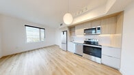 Located along Rue de la Commune in Montréal, this one-bedroom, one-bathroom unit offers a clean, modern design with light hardwood flooring and a crisp all-white palette throughout. The open-concept living and kitchen area is the heart of the unit, featuring a well-appointed kitchen with stainless steel appliances — including a refrigerator, electric range, dishwasher, and microwave — complemented by light wood cabinetry, open shelving, and a white tile backsplash. A large window floods the living area with natural light and frames views of the surrounding cityscape. The bedroom is a quiet, well-lit space with its own window offering expansive outdoor views, and includes a built-in closet with a sliding door for efficient storage. The bathroom is finished with large-format floor tiles and features a full soaking tub with an overhead shower. A dedicated hallway connects all spaces and houses a neatly integrated in-unit stacked washer and dryer within a discreet built-in closet — a practical convenience rarely found in similarly sized units. The overall ambiance is contemporary and functional, with consistent finishes that give the unit a cohesive, move-in-ready feel.
