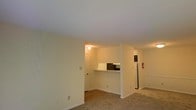 This single-floor space showcases an open-concept design that connects the living area to the kitchen through a convenient pass-through counter. The living area is finished with wall-to-wall carpeting in a neutral tone, contributing to a warm and cohesive aesthetic. Cream-colored walls and a smooth white ceiling create a bright, clean backdrop throughout the space. The kitchen features dark cabinetry visible beyond the pass-through counter, and a full-size refrigerator is positioned along the far wall, offering practical functionality. A doorway visible from the living area leads to additional spaces within the unit, while the open sightlines between rooms enhance the overall sense of space. Overhead lighting provides consistent illumination throughout. White baseboards frame the perimeter of the room, adding a crisp finishing detail to the neutral palette. The layout is straightforward and functional, making efficient use of the available floor plan while maintaining an uncluttered, move-in-ready appearance.