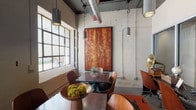 This single-floor business center showcases a compelling industrial-modern aesthetic throughout its expansive open layout. The architectural design retains raw, warehouse-inspired elements including exposed ductwork, structural columns, and high ceilings fitted with skylights that flood the space with abundant natural light. Pendant lighting and track fixtures complement the natural illumination, creating a dynamic and well-lit environment.

The space is organized into distinct functional zones. A dining and meeting area features dark wood tables paired with mid-century modern chairs, anchored by a large, striking abstract painting in warm amber and rust tones mounted against a textured concrete wall. Multi-pane industrial windows provide outdoor views and additional natural light. Decorative accents, including a vintage gramophone and sculptural tabletop pieces, add character.

A separate lounge area offers generous sectional seating in rich terracotta-toned upholstery arranged around low metal side tables, creating an inviting setting for informal gatherings. A large projection screen mounted on a painted masonry wall, flanked by colorful abstract artwork, serves as a focal point for presentations or screenings. Wood-paneled partitions define zones while maintaining the open, connected feel. The overall ambiance is creative, eclectic, and professionally welcoming.