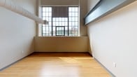 This two-floor loft-style space blends industrial character with functional living. The kitchen is well-appointed with warm wood cabinetry, granite countertops, light hardwood flooring, and a full suite of stainless steel appliances including a range, microwave, dishwasher, and refrigerator. Exposed ductwork runs along the upper portion of the kitchen, reinforcing the industrial aesthetic that carries throughout the space.

The living and bedroom areas feature neutral-toned carpeting, painted exposed ceiling beams, and ceiling fans that contribute to both the loft character and air circulation. Generous closet space is provided in the bedroom areas, with built-in double closets offering ample storage. Large windows — including a striking multi-pane grid window on one floor — allow natural light to flow in and provide views of the surrounding urban streetscape.

The space includes two bathrooms: one with a walk-in shower stall equipped with grab bars and dark wood cabinetry with granite countertops, and another featuring a full tub-shower combination. Both bathrooms are finished in neutral tones with clean, functional fixtures.

Throughout both floors, the consistent use of neutral walls, exposed structural elements, and practical finishes creates a cohesive, urban industrial ambiance suited for comfortable everyday living.