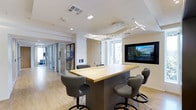 This single-floor space showcases a thoughtfully designed interior that blends functionality with modern aesthetics. The central feature is a sleek, light wood and dark-base high-top table surrounded by cushioned gray ergonomic bar-height chairs, creating an inviting collaborative hub. A striking sculptural light fixture hangs overhead, featuring an artistic branching design with integrated linear LED elements that serve as both a functional and decorative focal point.

Natural light filters in through windows along the wall, complementing the warm wood-toned flooring that runs throughout the space. A wall-mounted flat-screen display is prominently positioned, suggesting the area is equipped for presentations or media use. Neutral white walls and clean architectural lines contribute to a crisp, professional atmosphere.

Looking beyond the central work area, a corridor extends toward glass-partitioned rooms, offering a glimpse of additional private or semi-private spaces. Artwork adorns the walls near the corridor entrance, adding a curated, polished character to the overall environment. The combination of natural materials, contemporary lighting, and integrated technology makes this a well-appointed, versatile space.