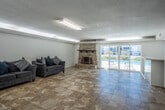 Located at 20455 Anza Ave in Torrance, California, this two-floor recreational space offers a well-rounded layout designed for communal gathering and leisure. The ground floor is generously proportioned, featuring warm travertine tile flooring throughout and a standout stacked-stone fireplace that anchors the main lounge area. Comfortable seating arrangements make it ideal for relaxed social gatherings, while sliding glass doors open to a view of an outdoor pool, filling the space with natural light and a seamless indoor-outdoor connection. Also on the ground floor is a kitchenette area equipped with refrigerators and counter space, providing practical functionality alongside a staircase with a sleek black metal railing that leads to the upper level. The upper floor transitions to rich hardwood flooring and features coffered ceiling details with exposed beams that lend architectural character to the space. Arched windows line the walls, allowing abundant natural light to pour in while offering views of the surrounding property. This level is well-suited as a game or activity room, with open floor space and an open-railing overlook down to the ground floor, creating a connected, airy feel between the two levels. The textured ceilings are consistent throughout, adding a classic finish to both floors.