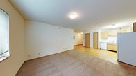 This single-floor unit features an open-concept layout that seamlessly connects the living area to the kitchen and additional rooms. The living space is finished with wall-to-wall carpeting in a warm neutral tone, providing a comfortable and inviting atmosphere. A flush-mount ceiling light illuminates the room, which is framed by clean white walls and standard baseboards.

Transitioning toward the back of the unit, the flooring shifts to smooth hardwood, leading to what appears to be a kitchen area outfitted with light wood cabinetry and a white appliance, likely a dishwasher or refrigerator. Two interior doors with medium wood-toned finishes are visible, suggesting access to additional rooms or closet storage. An electrical panel is mounted on the wall nearby, adding a practical element to the space.

The textured ceiling and consistent neutral palette throughout lend the unit a clean, functional aesthetic. The open flow between the living and kitchen areas makes the overall layout feel spacious and practical, suitable for everyday living.
