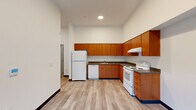 This single-floor unit offers three bedrooms, one bathroom, and a functional layout designed to maximize every square foot of its modest footprint. The kitchen is a standout feature, showcasing warm medium-toned wood cabinetry arranged in an L-shaped configuration with ample upper and lower storage. Dark laminate countertops provide contrast, while white appliances — including a refrigerator, electric range, and dishwasher — offer full functionality. Recessed lighting and tall ceilings give the kitchen an open, airy feel. Light wood-style luxury vinyl flooring runs consistently throughout the unit, complemented by crisp white walls and dark baseboards that lend a clean, contemporary aesthetic. The bedrooms are bright and simply finished, each featuring windows that bring in natural light and views of the surrounding exterior. The primary bedroom includes a closet with built-in storage. A connecting hallway links the rooms efficiently. The bathroom features a warm wood vanity with gray countertop, a medicine cabinet mirror, and well-placed wall lighting. The unit's clean, minimalist design and consistent material palette throughout make it a practical and functional living space.