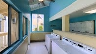 This shared laundry facility makes a memorable impression with its bold teal and soft yellow color scheme, creating a lively and welcoming atmosphere. The space features soaring vaulted ceilings with exposed white-painted beams, adding a sense of openness and architectural character uncommon in utility spaces. Overhead, a ceiling fan and a classic pendant light provide both airflow and warm illumination, complementing the natural light that pours in through the large windows. The windows overlook a lush outdoor area, offering a pleasant view and further brightening the interior. Multiple top-loading and front-loading washers and dryers are neatly arranged along the countertop walls, providing generous capacity for residents. The light-tiled flooring is clean and easy to maintain, while a playful wall decal reading "Laundry: Loads of Loads of Fun" adds a touch of humor and personality to the space. The layout is thoughtfully organized to allow comfortable movement between machines. Overall, this laundry room combines practicality with an upbeat design aesthetic, making routine laundry tasks a more enjoyable experience.