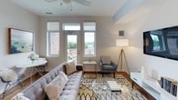 This thoughtfully designed single-floor residence offers a bright and contemporary living experience. The living room is welcoming and well-lit, featuring large windows that flood the space with natural light and provide outdoor views of the surrounding neighborhood. A ceiling fan adds a functional touch, while warm hardwood flooring anchors the room's modern aesthetic.

The kitchen is a standout feature, outfitted with crisp white cabinetry, open shelving, and a classic white subway tile backsplash. Stainless steel appliances — including a refrigerator, range, microwave, and dishwasher — are paired with sleek dark countertops and natural wood flooring, creating a clean and functional workspace ideal for cooking and entertaining.

The unit includes two bedrooms, both finished with soft carpet flooring and neutral tones. One bedroom benefits from dual windows with expansive views of the city skyline, while the other offers a quieter, more intimate setting. Both rooms are generously proportioned and well-suited for restful living.

The bathroom features a wide vanity with a stone-finish countertop, an under-mount sink, a large wall-spanning mirror, and elegant pendant lighting. A full tub and shower combination with a curtained surround completes the space. Throughout the unit, the consistent use of neutral palettes and modern fixtures creates a cohesive and inviting ambiance.