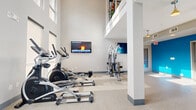 Located at 3902 W Tyvola Rd in Charlotte, NC, this well-appointed fitness center offers a comprehensive workout environment designed for both functionality and comfort. The space is characterized by impressively high ceilings that create an open, airy atmosphere, complemented by tall windows that allow abundant natural light to fill the room throughout the day.

The main exercise area is generously proportioned and houses a full array of cardio equipment, including treadmills, elliptical trainers, a recumbent bike, and rowing machines from brands such as Spirit and True. A dedicated strength training zone features a multi-station cable machine and a free weights area with a dumbbell rack and adjustable bench. Wall-mounted flat-screen televisions are positioned throughout for entertainment during workouts.

A standout feature of the space is a vibrant blue accent wall that adds a bold pop of color, complemented by matching blue cabinetry and built-in shelving. This area also includes a wall-mounted water station with a bottle-filling feature, storage cabinets, and a large framed mirror — providing a practical and refreshing corner for post-workout recovery. The speckled epoxy flooring throughout is both durable and easy to maintain. Pendant lighting adds a warm finishing touch to the space, creating an inviting and energizing environment for all fitness levels.