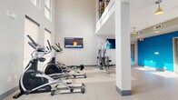 Located at 3902 W Tyvola Rd in Charlotte, NC, this well-appointed fitness center offers a comprehensive workout environment designed for both functionality and comfort. The space is characterized by impressively high ceilings that create an open, airy atmosphere, complemented by tall windows that allow abundant natural light to fill the room throughout the day.

The main exercise area is generously proportioned and houses a full array of cardio equipment, including treadmills, elliptical trainers, a recumbent bike, and rowing machines from brands such as Spirit and True. A dedicated strength training zone features a multi-station cable machine and a free weights area with a dumbbell rack and adjustable bench. Wall-mounted flat-screen televisions are positioned throughout for entertainment during workouts.

A standout feature of the space is a vibrant blue accent wall that adds a bold pop of color, complemented by matching blue cabinetry and built-in shelving. This area also includes a wall-mounted water station with a bottle-filling feature, storage cabinets, and a large framed mirror — providing a practical and refreshing corner for post-workout recovery. The speckled epoxy flooring throughout is both durable and easy to maintain. Pendant lighting adds a warm finishing touch to the space, creating an inviting and energizing environment for all fitness levels.