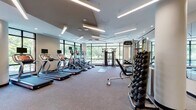 This contemporary fitness center is thoughtfully designed to support a comprehensive workout experience. The main exercise area is generously proportioned, featuring a row of Star Trac treadmills and elliptical machines aligned along expansive floor-to-ceiling glass windows that flood the space with natural light and offer pleasant views of surrounding greenery and the urban landscape beyond. A dedicated strength training zone includes a Life Fitness cable machine, adjustable benches, and a full rack of hex dumbbells mounted along the wall, providing a wide range of resistance training options. A separate cycling studio features spin bikes and stability balls, set against its own wall of large windows that frame outdoor views, creating an energizing and open atmosphere. The flooring transitions between plush carpet in the main workout area and hardwood in the cycling room, offering both comfort and functionality. The ceiling design incorporates geometric recessed panels with linear LED lighting that adds a modern, polished aesthetic throughout. A water station and entry vestibule with a decorative patterned rug complete the space, ensuring convenience and a welcoming arrival experience. Full-length mirrors in the strength area enhance the sense of space and allow for proper form monitoring during workouts.