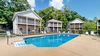 The outdoor pool area at Savannah Square Apartments in Auburn, Alabama, serves as a central communal amenity within the residential community. The generously sized swimming pool features clear blue water and a geometric design, making it a prominent focal point of the outdoor space. A wide concrete sundeck surrounds the pool on multiple sides, providing ample room for lounging and recreation. White lounge chairs are positioned throughout the deck, offering residents comfortable spaces to relax and enjoy the outdoors.

The pool area is enclosed by a chain-link fence lined with well-maintained shrubs, providing both security and a degree of privacy. Mature trees frame the perimeter of the space, contributing natural shade and a lush, green backdrop that enhances the overall ambiance. A lamp post is visible near the pool deck, indicating the area is equipped for evening use.

Surrounding the pool on multiple sides are two-story residential buildings characterized by traditional Southern architectural styling, featuring covered balconies, colonial-style columns, and neutral exterior siding in shades of gray and blue. The combination of thoughtful landscaping, communal seating, and the well-maintained pool creates an inviting and functional outdoor retreat within the community.