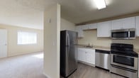 This single-floor space offers a clean, functional layout designed for comfortable everyday living. The kitchen is a standout feature, equipped with stainless steel appliances including a full-size refrigerator and dishwasher. White cabinetry provides ample storage, while light stone-toned countertops and a single-basin sink add to the kitchen's practical appeal. Wood-look flooring runs throughout the kitchen area, offering durability and easy maintenance. A textured ceiling adds subtle visual character throughout the space. Adjacent to the kitchen, the living area transitions seamlessly via an open-plan layout, featuring plush carpeting that adds warmth and comfort underfoot. A window in the living area allows natural light to filter in, creating a bright and welcoming atmosphere. The neutral wall tones throughout the space provide a versatile backdrop, making it easy to personalize with furnishings and décor. Overall, the space strikes a balance between practicality and comfort, with a cohesive design that feels both modern and approachable.