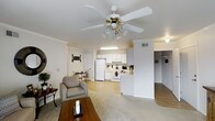 This single-floor residence is designed around an open-concept floor plan that seamlessly connects the living room, dining area, and kitchen into one cohesive living space. The living room serves as the central hub, featuring plush carpeted flooring, crown molding, and a ceiling fan with an integrated light fixture that provides both comfort and illumination. A wood-framed coffee table and cushioned armchair anchor the space, complemented by subtle decorative accents.

Adjacent to the living area, a small dining nook accommodates a compact table and chairs, ideal for casual meals. The kitchen is situated toward the rear of the open layout and is outfitted with white cabinetry, white appliances including a refrigerator and range, and a peninsula that provides a natural boundary between the kitchen and living spaces. Light-colored countertops and a flush ceiling light keep the kitchen bright and functional.

The flooring transitions from carpet in the living and dining areas to hard surface flooring in the kitchen. A hallway visible beyond the living area suggests additional rooms. The overall palette of whites and warm neutrals gives the space a clean, welcoming ambiance throughout.