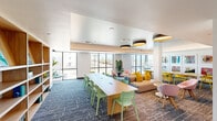 This single-floor communal space is a thoughtfully designed lounge and co-working area that seamlessly blends social and functional zones into one cohesive, energetic environment. The layout is anchored by a long, solid wood communal table lined with brightly colored chairs — mint green on one side, vivid yellow on the other — equipped with built-in power access points for convenience. The dining and work area is framed by a striking gallery wall of bold, large-format contemporary artwork, adding a vibrant artistic identity to the space. Warm wood built-in shelving lines the back wall, offering both decorative display and practical storage.

The living and lounge area features a neutral sectional sofa accented with yellow throw pillows, complemented by playful pink accent chairs and small side tables, creating an inviting space for relaxed conversation or casual gatherings. Floor-to-ceiling windows along the perimeter flood the interior with natural light and offer open views of the surrounding landscape, including palm trees and adjacent architecture.

Ceiling details vary throughout, with tray ceilings, a sky-blue painted ceiling section, and large drum pendant light fixtures adding visual depth and dimension. Patterned carpet tiles in blue and brown tones ground the space, while curated artwork — including large abstract canvases near the entry corridor — reinforces the space's creative and contemporary character throughout.