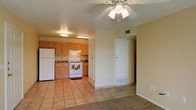 This single-story space is designed with an open-concept layout that connects the living area to the kitchen, creating a cohesive and functional environment. The living area features neutral carpet flooring and warm yellow-toned walls, contributing to a welcoming ambiance. A ceiling fan with a built-in light fixture is centrally mounted, providing both air circulation and ambient lighting to the space.

The kitchen is positioned at the rear of the open area and is outfitted with light wood cabinetry offering ample storage. White appliances, including a gas range and a full-sized refrigerator, are complemented by a simple under-cabinet range hood. The kitchen flooring transitions to tan ceramic tile, which also extends toward the entryway. A double-basin sink is situated beneath the cabinetry along the back wall.

A white paneled door near the kitchen leads to an adjacent room or exterior, with a floor-level vent indicating central climate control throughout. The overall design is practical and straightforward, with consistent warm, neutral tones unifying the space.