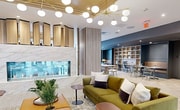 This expansive 3,520 square foot space showcases a harmonious blend of contemporary design and functional versatility. The open floor plan is anchored by a striking ceiling feature - a golden recessed panel adorned with an array of spherical pendant lights, creating a warm and inviting atmosphere.

The main area boasts a variety of seating options, from comfortable lounge sofas to practical dining tables and chairs, catering to both relaxation and work needs. A patterned tile floor adds visual interest, contrasting beautifully with the herringbone wood flooring in adjacent areas.

Large windows flood the space with natural light, offering views of the surrounding urban landscape. The kitchen area, visible through a glass partition, features sleek cabinetry and modern appliances. A central brick column adds a touch of industrial chic, while built-in shelving units provide ample storage and display space.

The color palette is predominantly neutral, with charcoal grey walls and warm wood tones, punctuated by pops of color in the form of green upholstery. High-tech amenities are seamlessly integrated, including a large screen display and various work stations.

Private meeting rooms and work pods are available for more focused tasks, while the main area encourages collaboration and social interaction. This thoughtfully designed space effortlessly balances professionalism with comfort, making it ideal for a modern, dynamic work environment.