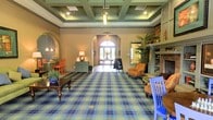 This single-floor space presents an elegantly designed lobby that blends traditional charm with a warm, welcoming ambiance. The room is defined by its striking coffered ceiling, finished in sage green and trimmed with recessed lighting and a central pendant fixture that draws the eye upward and adds a sense of grandeur. Underfoot, a bold blue and green plaid carpet anchors the space with a distinctive, classic character.

Arched doorways frame the room beautifully, with a central arched entryway at the far end offering a view to the outdoors, flooding the interior with natural light. Additional arched alcoves on either side of the space add architectural depth and interest.

To one side, a fireplace with a decorative mantel and built-in shelving creates a cozy seating nook, furnished with upholstered armchairs and accented by lush tropical plants. On the opposite side, a casual dining or lounge area features a dark wood table accompanied by vibrant orange chairs, adding a pop of color to the warm, neutral walls.

A small console table with a table lamp and a chalkboard sign contribute to the space's approachable, community-oriented feel. Overall, the lobby exudes a comfortable, resort-like quality that is both functional and aesthetically refined.