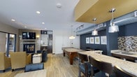 This single-floor space is designed as a stylish recreational and social lounge. The open layout accommodates multiple activity zones that flow seamlessly into one another. At the center of the space sits a full-size pool table beneath modern pendant lighting, serving as the focal point of the room. Track lighting installed along the ceiling supplements the ambient recessed lighting throughout.

To one side, a bar-height counter with granite or stone-patterned surfaces provides casual seating with sleek dark chairs, ideal for socializing or casual dining. A lounge area is situated toward the back of the space, featuring comfortable seating arranged around a wall-mounted flat-screen television and a decorative fireplace with textured tile surround, creating an inviting gathering spot.

The walls are finished in a deep navy blue accent color, complemented by framed artwork that adds a polished touch. Exposed ductwork along the ceiling reinforces an industrial aesthetic, while light wood-toned flooring runs throughout, brightening the space. The combination of functional recreational features and contemporary design makes this a well-appointed communal entertainment area.