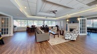 Located at 2400 Milhaven Dr in Henrico, Virginia, this single-story commercial space combines a welcoming, residential-inspired aesthetic with a fully functional professional environment. The open-concept layout flows seamlessly from a comfortable reception and lounge area into a dedicated workstation zone, creating a versatile setting suited for client-facing operations.

The lounge area serves as an inviting focal point, furnished with upholstered sofas and accent chairs arranged around a glass-top coffee table atop a neutral area rug. A white-mantled fireplace with a decorative surround anchors one wall beneath soft blue accent paint, contributing warmth and character. Wall sconces flank the fireplace, adding an inviting glow.

Adjacent to the lounge, the open office area accommodates multiple workstations positioned along expansive windows that span nearly the full width of the room. These windows flood the space with natural light and provide views of the surrounding greenery. The raised tray ceiling with recessed lighting and a ceiling fan enhances the sense of volume overhead, while wood-tone flooring ties the entire space together with a clean, polished finish. French doors connect to additional areas, offering flexibility in layout and privacy.