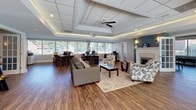 Located at 2400 Milhaven Dr in Henrico, Virginia, this single-story commercial space combines a welcoming, residential-inspired aesthetic with a fully functional professional environment. The open-concept layout flows seamlessly from a comfortable reception and lounge area into a dedicated workstation zone, creating a versatile setting suited for client-facing operations.

The lounge area serves as an inviting focal point, furnished with upholstered sofas and accent chairs arranged around a glass-top coffee table atop a neutral area rug. A white-mantled fireplace with a decorative surround anchors one wall beneath soft blue accent paint, contributing warmth and character. Wall sconces flank the fireplace, adding an inviting glow.

Adjacent to the lounge, the open office area accommodates multiple workstations positioned along expansive windows that span nearly the full width of the room. These windows flood the space with natural light and provide views of the surrounding greenery. The raised tray ceiling with recessed lighting and a ceiling fan enhances the sense of volume overhead, while wood-tone flooring ties the entire space together with a clean, polished finish. French doors connect to additional areas, offering flexibility in layout and privacy.