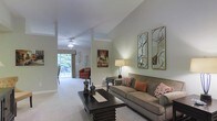 This single-story space showcases a thoughtfully designed living area highlighted by soaring vaulted ceilings that lend an expansive, airy feel to the interior. The living room is finished in soft sage green tones, creating a calm and welcoming ambiance throughout. Plush, wall-to-wall carpeting in a neutral hue runs underfoot, adding warmth and comfort to the space.

A generously sized taupe sofa serves as the centerpiece of the living area, complemented by accent pillows in rich burnt orange and patterned fabrics that add depth and visual interest. A substantial dark wood coffee table anchors the seating arrangement, offering both style and function. Two decorative gold-framed circular mirrors adorn the wall, enhancing the sense of light and sophistication.

A ceiling fan is installed overhead, contributing to the practicality of the space. At the far end of the room, a large window dressed with valance curtains offers views of lush greenery outside, flooding the area with natural light. A cozy accent chair and additional wall art complete the composition, giving the space a polished, well-curated character.