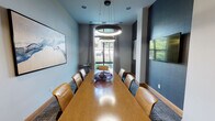 This sophisticated conference room is designed to facilitate productive meetings in a refined, professional environment. At its center sits a long, warm-toned wood conference table surrounded by tan leather chairs, providing ample seating for a sizable group. Two striking circular ring pendant lights hang overhead, casting a focused, modern glow across the table's polished surface.

The room's color palette contrasts soft light gray walls with a deep charcoal accent wall, creating a dynamic yet professional atmosphere. A wall-mounted flat-screen television is positioned on the darker accent wall, making it ideal for presentations. Complementing the space are several curated abstract art pieces that add a creative dimension to the interior.

Natural light enters through large windows draped with neutral linen curtains, offering views of greenery outside. A utility cart near the window provides a convenient surface for materials or refreshments. The floor is covered in a textured, patterned carpet that adds visual depth underfoot.

A glass-paneled sliding door provides entry to the room, maintaining an open, transparent feel while offering separation from adjacent spaces. The overall ambiance is polished and contemporary, balancing functionality with thoughtful design details.