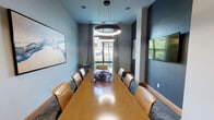 This sophisticated conference room is designed to facilitate productive meetings in a refined, professional environment. At its center sits a long, warm-toned wood conference table surrounded by tan leather chairs, providing ample seating for a sizable group. Two striking circular ring pendant lights hang overhead, casting a focused, modern glow across the table's polished surface.

The room's color palette contrasts soft light gray walls with a deep charcoal accent wall, creating a dynamic yet professional atmosphere. A wall-mounted flat-screen television is positioned on the darker accent wall, making it ideal for presentations. Complementing the space are several curated abstract art pieces that add a creative dimension to the interior.

Natural light enters through large windows draped with neutral linen curtains, offering views of greenery outside. A utility cart near the window provides a convenient surface for materials or refreshments. The floor is covered in a textured, patterned carpet that adds visual depth underfoot.

A glass-paneled sliding door provides entry to the room, maintaining an open, transparent feel while offering separation from adjacent spaces. The overall ambiance is polished and contemporary, balancing functionality with thoughtful design details.