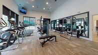 This modern fitness center is thoughtfully designed to support a full range of workout routines across two distinct zones. The main gym area features soaring ceilings and large windows that flood the space with natural light while offering views of the surrounding outdoor amenities. The strength training section is outfitted with a comprehensive selection of cable machines, squat racks, a full dumbbell rack, and adjustable benches, all positioned along a floor-to-ceiling mirror wall that enhances the sense of openness. The cardio zone lines the windowed wall, housing stair climbers, ellipticals, and a recumbent bike, all facing the outdoors for an energizing workout experience. Wall-mounted televisions are conveniently placed throughout. A separate spin and group fitness studio provides an intimate, high-energy environment with multiple stationary cycling bikes arranged facing expansive windows. This studio also features a striking exposed brick accent wall adorned with motivational typography, adding character and energy to the space. A well-organized equipment rack holds a variety of exercise balls, foam rollers, resistance bands, and kettlebells. Durable wood-look flooring in the studio and plush carpet in the main gym area offer both functionality and comfort throughout.
