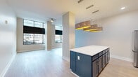 Located at 265 E State St in the heart of downtown Columbus, Ohio, this one-bedroom, one-bathroom residence offers a well-designed layout with modern finishes throughout. The open-concept living and kitchen area is a standout, anchored by a striking kitchen featuring navy blue shaker-style cabinetry, a large center island with a crisp white countertop, stainless steel appliances, and a subway tile backsplash. Pendant lighting with gold-accented drum shades adds a warm, contemporary touch above the island. The living area extends toward a wall of floor-to-ceiling windows fitted with blackout roller shades, flooding the space with natural light and offering direct views of the downtown streetscape. Light wood-tone flooring runs throughout the main living areas, complementing the clean, neutral palette. A ceiling fan provides additional comfort in the living room. The bedroom features soft carpeting, a window for natural light, and access to a dedicated closet. The bathroom is well-appointed with a glass-enclosed walk-in shower with tile surround, a navy vanity with a white countertop, and built-in open shelving for storage. A hallway connects the living spaces to the private areas, completing this thoughtfully arranged urban residence.