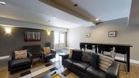 Located at 1600 West Lane Avenue in Upper Arlington, Ohio, this single-floor space offers a thoughtfully designed interior that balances comfort and functionality. The main living area features a bold, geometric black-and-white patterned floor that anchors the room and adds a striking visual element. A leather sofa and coordinating accent chairs — dressed with decorative pillows in warm gold and plaid tones — create an inviting seating arrangement centered around a coffee table. Recessed lighting is installed throughout the ceiling, complemented by wall-mounted sconces that provide warm ambient illumination. Structural ceiling beams add architectural character to the space. A wall-mounted piece of art depicting a forest scene introduces a natural, earthy touch to the neutral gray walls. Toward the rear of the space, a bar-height dining table with black stools serves as a defined area for dining or casual gathering. A windowed door at the back of the room allows natural light to filter in, brightening the space. The overall design is modern and cohesive, with a neutral color palette accented by warm tones, making it a versatile and welcoming environment.