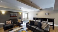 Located at 1600 West Lane Avenue in Upper Arlington, Ohio, this single-floor space offers a thoughtfully designed interior that balances comfort and functionality. The main living area features a bold, geometric black-and-white patterned floor that anchors the room and adds a striking visual element. A leather sofa and coordinating accent chairs — dressed with decorative pillows in warm gold and plaid tones — create an inviting seating arrangement centered around a coffee table. Recessed lighting is installed throughout the ceiling, complemented by wall-mounted sconces that provide warm ambient illumination. Structural ceiling beams add architectural character to the space. A wall-mounted piece of art depicting a forest scene introduces a natural, earthy touch to the neutral gray walls. Toward the rear of the space, a bar-height dining table with black stools serves as a defined area for dining or casual gathering. A windowed door at the back of the room allows natural light to filter in, brightening the space. The overall design is modern and cohesive, with a neutral color palette accented by warm tones, making it a versatile and welcoming environment.