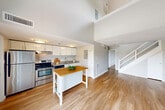 Located at 73 Charles St in Providence, RI, this two-bedroom, two-bathroom two-story unit offers a functional and well-appointed layout across two floors. The main level houses a bright, modern kitchen featuring sleek white cabinetry, stainless steel appliances including a refrigerator and range, a double sink, and warm hardwood flooring throughout. A center island with a butcher block top adds additional prep and dining space. Both bathrooms are clean and simply designed, each featuring a tub-shower combination, an undermount sink with a dark countertop vanity, and tile flooring. The upper level is a standout, boasting vaulted ceilings with dramatic angles, a skylight that floods the space with natural light, and expansive windows offering views of the Providence skyline and surrounding greenery. The generously sized primary bedroom continues the vaulted ceiling aesthetic and is complemented by multiple closets. The second bedroom on the upper floor also features angled ceilings and ample closet storage. Hardwood flooring flows throughout both levels, creating a cohesive look. A private balcony extends the living space outdoors, providing open views of the city and neighboring architecture.