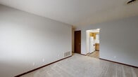 Located at 250 N East St in Plainfield, Indiana, this single-floor, three-bedroom, one-bathroom residence offers a practical and straightforward layout. The living area features neutral-toned carpet, light gray walls, and a textured ceiling, creating a clean and understated atmosphere. A large window allows natural light to fill the space and provides views of the surrounding neighborhood.

The kitchen is efficiently designed with warm wood cabinetry, a marble-patterned countertop, white appliances including a refrigerator and electric range with overhead ventilation, and a utility sink positioned beneath a window. The hard surface flooring in the kitchen adds durability and ease of maintenance. An adjacent area with additional cabinetry, countertop space, a pendant light, and a second sink provides extra functional workspace.

Each of the three bedrooms is carpeted and features dark wood-trimmed closets with bi-fold doors, offering ample storage. The primary bedroom is notably spacious and includes a large closet. The full bathroom is clean and simply appointed, featuring a tiled tub-shower combination with chrome fixtures. A utility room rounds out the floor plan, providing additional functional space. Dark wood door trim and baseboards are consistent throughout, lending a cohesive character to the home.