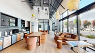 This thoughtfully designed single-floor space in Oakland, CA (94611) offers a generous open-plan layout that blends contemporary industrial architecture with warm, curated interiors. Soaring ceilings with exposed ductwork and structural steel lend an airy, loft-like quality to the space, while an eclectic array of pendant lighting in varied shapes and warm tones adds visual interest overhead.

Floor-to-ceiling black-framed glass windows line the exterior walls, flooding the interior with abundant natural light and providing unobstructed street-level views of the surrounding urban landscape. Light-toned hardwood flooring runs throughout, contributing to the bright and cohesive atmosphere.

The open main area is divided into distinct functional zones. A communal dining table with curved upholstered chairs anchors a central gathering space, while a lounge area features a sculptural leather sectional sofa alongside additional accent seating in rich terracotta tones. A well-appointed kitchenette incorporates green marble countertops, multi-toned cabinetry in blue, peach, and charcoal, a grey subway tile backsplash, and open shelving.

A glass-enclosed private room, visible from the main space, features dramatic floor-to-ceiling bookshelf wall graphics and a sleek built-in workspace, offering a quiet retreat within the larger open environment.