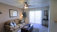 Located in Amarillo, Texas, this single-floor residence offers a warm and functional layout designed for comfortable everyday living. The living room serves as the heart of the space, featuring plush carpeted flooring, neutral-toned walls, and a textured ceiling fitted with a ceiling fan and light fixture — ideal for the Texas climate. A large sliding glass door floods the room with natural light and provides direct access to an outdoor area, giving the space an open, airy feel. Vertical blinds offer privacy and light control as needed. The living area is well-proportioned, accommodating a full sofa and a substantial coffee table, with room for decorative touches such as framed wall art and indoor plants. A floor lamp adds warm ambient lighting, enhancing the inviting atmosphere. The carpet and neutral color palette throughout create a cohesive and relaxed aesthetic. This single-story layout provides easy accessibility and a straightforward flow, making it a practical and welcoming home in the heart of Amarillo.