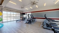Located at 7660 S Winnipeg St in Aurora, Colorado, this well-equipped fitness center is designed to support a full range of workout routines in a bright, energizing environment. The single-floor space features light wood-style flooring throughout and a bold red and gray horizontal stripe motif along the walls, giving the room a dynamic, athletic aesthetic. Ceiling fans and recessed fluorescent lighting ensure comfortable conditions during any workout.

The cardio zone is well-stocked with a row of elliptical trainers, stair climbers, and a treadmill, all positioned to take advantage of the natural light pouring through the large garage-style roll-up windows. These expansive windows open the space visually to the outdoors and bring in generous amounts of sunlight.

The strength and free-weight area includes multiple tiers of dumbbells on organized racks, a weight bench, and rubber floor mats for safety. A multi-function pull-up and dip station rounds out the strength training options. Wall-mounted mirrors expand the sense of space and allow for form monitoring during exercise.

A dedicated stretching and functional fitness area features stability balls, resistance tools, a yoga mat station, and open floor space. Mounted televisions are positioned throughout for entertainment. A small refreshment counter and mini-fridge add convenience, making this a comprehensive, amenity-rich facility.