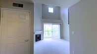 This two-floor residence is defined by its impressive vertical space and bright, open atmosphere. The living area immediately captures attention with its dramatically high ceilings and a clerestory window positioned near the roofline, flooding the room with natural light from above. Large sliding glass doors with vertical blinds open to an outdoor balcony, offering a view of surrounding greenery and further enhancing the sense of openness. A classic fireplace serves as a central focal point in the living area, adding warmth and character to the space. The flooring throughout the main living area is finished in plush, light-toned carpeting that contributes to the overall soft and neutral aesthetic. The walls are painted in a clean, contemporary grey-white palette that reflects light and keeps the space feeling airy and modern. A dedicated entryway with a full-sized door provides a clear separation between the interior and exterior. The two-floor layout offers distinct living zones, allowing for flexible use of space across levels. Overall, the residence combines functional design with architectural detail, creating a comfortable and well-lit environment suitable for everyday living.