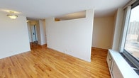 This single-floor residence presents a bright, open layout highlighted by warm-toned hardwood floors that run throughout the living area, creating a cohesive and inviting atmosphere. The main living space is generously proportioned, with smooth white ceilings and crisp neutral walls that provide a clean, move-in-ready canvas. A partial dividing wall with a recessed upper section adds subtle architectural interest while helping to define distinct zones within the open floor plan without fully closing off the space. Recessed outlets and fixtures are thoughtfully positioned along the baseboards, contributing to a tidy, uncluttered aesthetic. A glimpse into the adjoining areas reveals a mirrored sliding door — likely a closet — along with a white interior door, suggesting well-integrated storage and room connectivity. The light-filled, airy quality of the space is enhanced by the reflective sheen of the hardwood floors and the uniformity of the pale wall tones. Overall, the residence offers a functional and adaptable layout suited to a variety of living arrangements, with classic finishes that complement both modern and traditional décor styles.