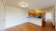 This single-floor residence offers a well-designed layout that balances functionality with modern finishes. The open-concept living and kitchen area serves as the heart of the space, where warm hardwood-style flooring extends seamlessly throughout, creating a cohesive and inviting atmosphere.

The kitchen is thoughtfully appointed with medium-toned wood cabinetry offering generous storage, granite countertops, and a matching granite tile backsplash that adds texture and visual interest. A peninsula-style counter extends into the living area, providing a practical breakfast bar that enhances the open flow between spaces. Black appliances, including a range, microwave, and dishwasher, complement the cabinetry, and a track lighting fixture ensures the kitchen is well-lit. A sink with a matte black faucet is positioned along the counter facing the living area.

The living area is bright and spacious, with a sliding glass door leading to a private balcony that overlooks a green outdoor setting. The bedrooms are calm and neutral, featuring the same hardwood-style flooring, windows with natural light, and access to walk-in closet space. The bathroom includes a full-sized soaking tub with a clean, all-white surround. Throughout the residence, clean white walls and a neutral palette create a versatile, move-in ready environment.