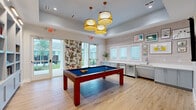 Located at 5901 Myrtle Dr in Wildwood, Florida, this single-floor community game room is a well-appointed recreational space designed for social gatherings and leisure. The centerpiece of the room is a solid wood pool table with a blue felt surface, positioned beneath a cluster of three woven pendant lights that lend a warm, tropical ambiance to the space.

Light hardwood flooring runs throughout, complementing the room's bright and airy aesthetic. A tray ceiling adds architectural depth, while recessed lighting provides additional illumination alongside the statement pendant fixtures. Large glass French doors along one wall flood the space with natural light and offer direct access to an outdoor area, framing views of the surrounding neighborhood.

Built-in shelving units with a powder-blue interior line one wall, providing display and storage space alongside a wall-mounted flat-screen television and a pool cue rack. On the opposite wall, a full wet bar setup features light gray cabinetry, a white countertop, an undermount sink, a stainless steel refrigerator, and a beverage cooler — ideal for entertaining.

Decorative touches include a bold tropical botanical wall panel near the entrance, a curated gallery wall of framed artwork, and lush potted greenery, all contributing to the room's fresh, Florida-inspired character.