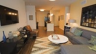 This single-floor residence showcases an open-concept layout that connects the living area and kitchen in one cohesive space. The living room is well-appointed, featuring a comfortable seating arrangement centered around a round white marble-top coffee table resting on a geometric patterned area rug. Warm hardwood flooring extends throughout, adding a sense of continuity and warmth across the space. A media console anchors one side of the living area, while a floor lamp and additional accent pieces contribute to the room's inviting ambiance.

The kitchen sits centrally within the open floor plan, highlighted by a white island with granite countertops that provides both workspace and a visual focal point. Stainless steel appliances, including a refrigerator, are visible, and pendant lighting above the island adds a stylish touch. White cabinetry and clean lines give the kitchen a modern, organized feel.

Additional rooms are accessible through doorways visible from the main living area, suggesting a practical and well-organized floor plan. The warm, neutral color palette used throughout creates a cohesive and welcoming atmosphere.