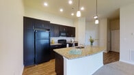 Located at 130 Ocean Aisle Circle in Salisbury, Maryland, this single-floor residence offers a thoughtfully designed living space with modern finishes throughout. The kitchen stands as a central highlight, showcasing rich dark espresso cabinetry that extends from floor to ceiling, providing generous storage. Striking granite countertops in warm gold and cream tones complement the cabinetry beautifully, while a spacious kitchen island with an undermount sink serves as both a functional workspace and a casual gathering spot. Black appliances, including a full-sized refrigerator, range, and built-in microwave, lend a sleek, cohesive aesthetic to the space. Warm hardwood-style flooring runs throughout the kitchen and adjoining areas, adding a sense of continuity and warmth. Recessed lighting and elegant pendant fixtures illuminate the kitchen, creating a bright and inviting atmosphere. The open layout connects the kitchen seamlessly to the surrounding living areas, enhancing the sense of space and flow. Neutral wall tones provide a versatile backdrop, allowing the bold cabinetry and countertop details to take center stage throughout this well-crafted, move-in-ready home.