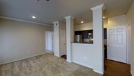 This single-floor space showcases an open-concept layout that seamlessly connects the living area to the kitchen. Decorative columns with white crown molding detailing serve as an architectural divider, adding visual interest while maintaining an open feel between the spaces. The living area features neutral-toned carpeting and warm beige walls, creating a welcoming atmosphere. Recessed lighting illuminates the space throughout.

The kitchen is defined by dark espresso cabinetry that contrasts elegantly with granite countertops. Stainless steel appliances, including a refrigerator, microwave, and range, lend a modern touch. The kitchen counter extends partially as a peninsula, offering a functional transition point between the cooking and living areas. The flooring in the kitchen transitions to a hard surface, distinguishing it from the carpeted living space.

Two white panel doors are visible toward the back of the unit, suggesting access to additional rooms or storage. The neutral color palette and consistent use of white trim throughout contribute to a cohesive, clean aesthetic.