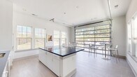 Located at 2525 Sycamore Farms Dr NW in Concord, NC, this single-floor community kitchen and dining space offers a sleek, modern design that seamlessly blends functionality with contemporary style. The open-concept layout centers around a striking central island topped with a bold, deep-blue countertop that anchors the room and serves as a natural gathering point. Crisp white cabinetry lines the walls, complemented by a matching blue countertop and backsplash along the perimeter, where a stainless steel sink, a wall-mounted television, and a built-in beverage cooler add to the room's utility and appeal. Large-format light gray tile flooring runs throughout, lending the space a clean, polished look. Natural light pours in generously through multiple windows fitted with white horizontal blinds, offering views of the surrounding greenery. A defining architectural feature is the full-width glass panel garage-style door, which opens the dining area to the outdoors, creating an airy indoor-outdoor connection. The dining area accommodates bistro-style high tables and chairs, ideal for casual gatherings. Colorful framed artwork adorns the walls, adding warmth and personality to the otherwise minimalist palette. The overall ambiance is bright, fresh, and inviting.