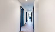 Located at 401 E State St in the heart of downtown Ithaca, NY, this two-bedroom, one-bathroom unit offers a thoughtfully designed single-floor layout with a contemporary industrial aesthetic. Light wood-tone flooring and crisp white walls flow consistently throughout, unified by bold navy blue door frames and baseboards that serve as a distinctive design signature.

The open-concept kitchen and living area forms the centerpiece of the unit. The kitchen is outfitted with slate-blue flat-front cabinetry, granite countertops, a subway tile backsplash, stainless steel appliances, and a matte black faucet. A substantial kitchen island with a white stone top and fluted base provides additional workspace and seating. Exposed ceiling beams and industrial-style pendant lighting add architectural character.

Both bedrooms are generously sized, each featuring expansive floor-to-ceiling, grid-paned windows that frame views of the downtown Ithaca skyline and flood the rooms with natural light. The primary bedroom is particularly spacious and well-lit.

The bathroom features large-format gray tile, a navy vanity with a framed mirror, and matte black fixtures. A long hallway connects all rooms efficiently, and the unit also includes dedicated laundry and two closets.