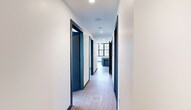 Located at 401 E State St in the heart of downtown Ithaca, NY, this two-bedroom, one-bathroom unit offers a thoughtfully designed single-floor layout with a contemporary industrial aesthetic. Light wood-tone flooring and crisp white walls flow consistently throughout, unified by bold navy blue door frames and baseboards that serve as a distinctive design signature.

The open-concept kitchen and living area forms the centerpiece of the unit. The kitchen is outfitted with slate-blue flat-front cabinetry, granite countertops, a subway tile backsplash, stainless steel appliances, and a matte black faucet. A substantial kitchen island with a white stone top and fluted base provides additional workspace and seating. Exposed ceiling beams and industrial-style pendant lighting add architectural character.

Both bedrooms are generously sized, each featuring expansive floor-to-ceiling, grid-paned windows that frame views of the downtown Ithaca skyline and flood the rooms with natural light. The primary bedroom is particularly spacious and well-lit.

The bathroom features large-format gray tile, a navy vanity with a framed mirror, and matte black fixtures. A long hallway connects all rooms efficiently, and the unit also includes dedicated laundry and two closets.