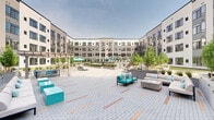 Located at 415 Stump Rd in Montgomeryville, Pennsylvania, this expansive outdoor courtyard amenity space is nestled within the U-shaped, multi-story modern building known as Immerse. The courtyard is defined by a contemporary architectural aesthetic, featuring a neutral palette of gray siding and large windows that frame the open-air environment on all sides.

The space is thoughtfully divided into several distinct activity zones. A central paved plaza features decorative brick-patterned pavers with colorful accent striping, providing a generous open gathering area. Comfortable outdoor lounge seating with teal accent cushions and a sleek fire pit table create an inviting social hub. Sun loungers and a pergola-shaded dining area offer relaxed spots for unwinding, while teal umbrellas provide shade near a resort-style pool deck.

Lush greenery is woven throughout, with tall bamboo, ornamental grasses, and manicured plantings housed in large raised planters that add natural texture and visual privacy between zones. A synthetic turf lawn area hosts recreational activities, including cornhole and a life-sized chess set on a dedicated deck, catering to an active, social lifestyle. Outdoor grilling stations complete the space, making it well-suited for communal gatherings. The overall ambiance is modern, vibrant, and resort-inspired.
