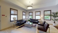This well-appointed single-floor communal space is designed with a contemporary aesthetic that balances comfort and sophistication. The lounge area features multiple seating arrangements composed of sleek black leather sofas, rich brown leather accent chairs with gold-toned frames, and a mirrored-metallic glass coffee table anchored on layered area rugs over light hardwood flooring. Abundant natural light filters through numerous windows fitted with warm wood trim and woven Roman shades, offering views of lush greenery outside. A large pendant drum light with a brushed finish serves as the central fixture, complemented by elegant floor and table lamps throughout. Potted tropical plants add vibrant, organic touches to the space. The adjoining dining and social area includes marble-topped dining tables with modern gray chairs, pendant lighting, and light wood flooring that flows seamlessly throughout. A built-in bar counter with dark wood cabinetry, bar-height seating, a wall-mounted television, and beverage stations make this zone ideal for gathering and entertaining. Glass doors provide direct access to an outdoor area, enhancing the open, airy ambiance of the space overall.
