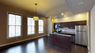This single-floor space showcases a thoughtfully designed open-concept layout that seamlessly connects the kitchen and dining areas. The kitchen is a standout feature, outfitted with rich dark cabinetry, granite countertops, and a full suite of stainless steel appliances including a range and over-the-range microwave. A generous kitchen island with a breakfast bar overhang provides additional prep space and casual seating opportunities. Recessed lighting illuminates the kitchen, complemented by a stylish geometric pendant light that anchors the dining area. Deep, dark-stained hardwood flooring flows continuously throughout the space, adding warmth and a sense of cohesion. Large windows along the exterior wall flood the dining area with natural light, enhancing the open and airy atmosphere. Neutral warm-toned walls provide a versatile backdrop that complements the bold cabinetry and flooring. The tray ceiling detail above the kitchen adds architectural interest and dimension to the space. Overall, the layout balances functionality with modern aesthetic appeal, making it well-suited for both everyday living and entertaining.